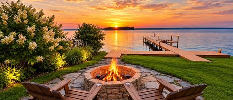 A fire pit with two wooden chairs, a stone fire pit, and a wooden dock extending into a lake at sunset.