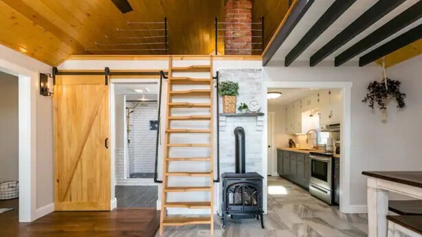 A spacious loft-style living area with a wooden floor, a loft bed, a fireplace, and a kitchen in the background. - Luxury Farm Cottage, Hot Tub, Sauna,Massage,Shenandoah Views, pet friendly (Weyers Cave)