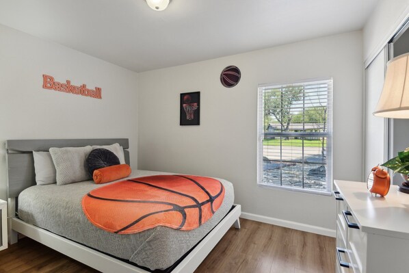 A bedroom with a bed, a basketball-themed bedspread, a basketball wall decal, a window with a view of the outdoors, and a bedside table with a lamp. - Cozy Rental Near Medical Center, UTSA, Six Flags, Lackland AFB, La Cantera, Rim (San Antonio)