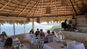 Un restaurante con techo de paja y vistas al océano. Las mesas están preparadas con manteles blancos y sillas.