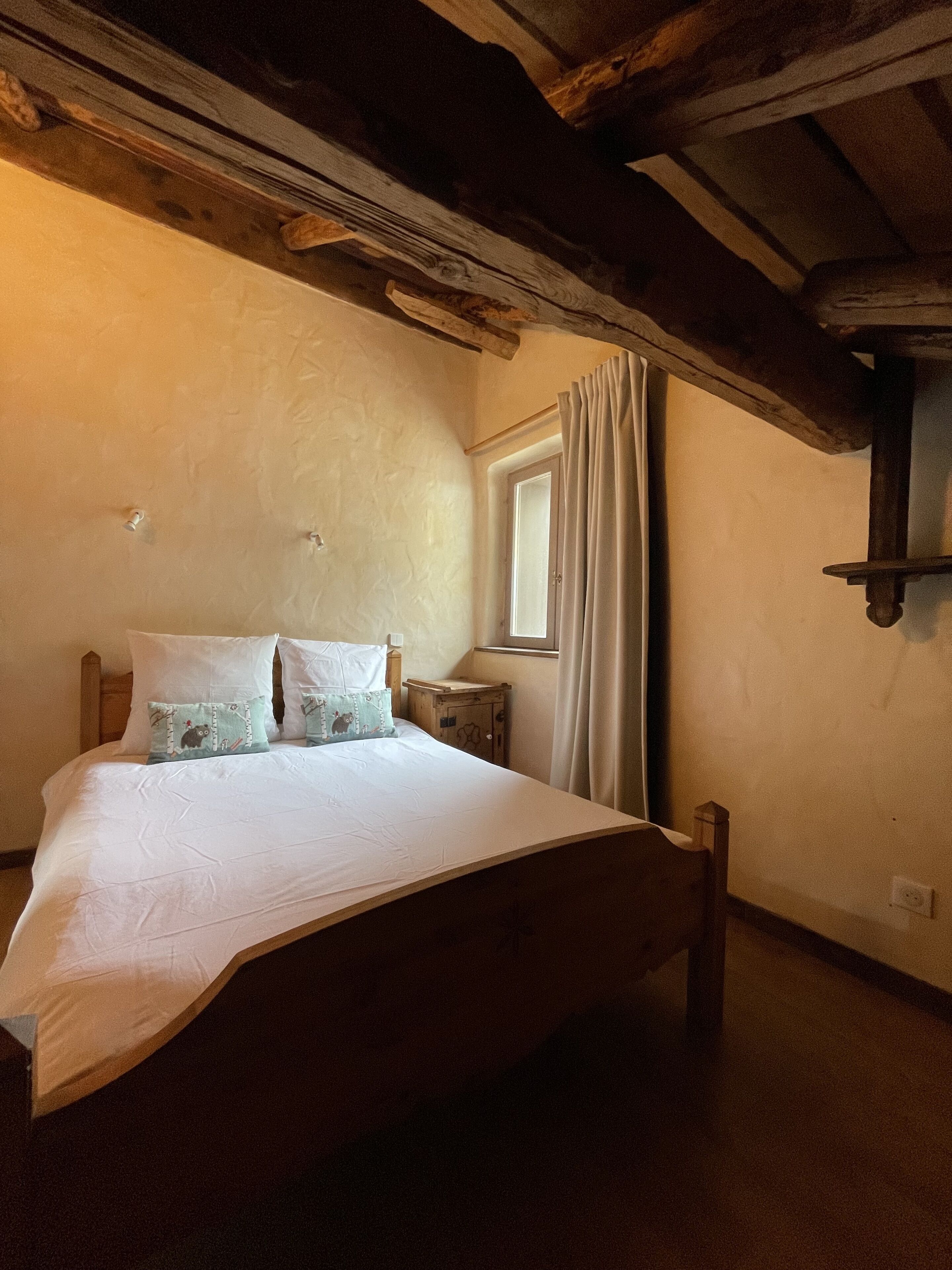 Un dormitorio con cama de madera, una mesita de noche, una ventana con cortinas y una viga de techo de madera.