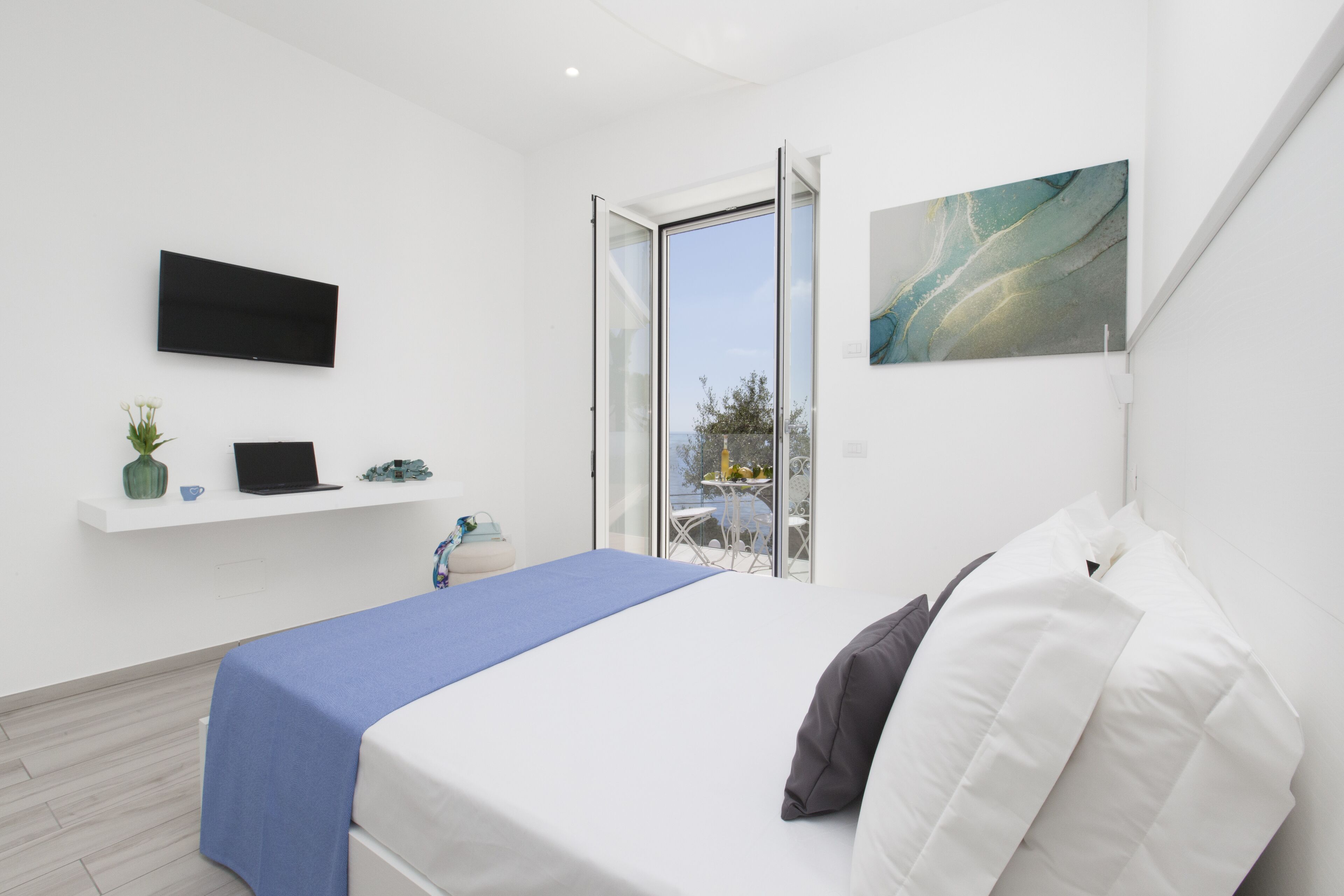 Ein modernes Schlafzimmer mit Bett, einem an der Wand montierten Fernseher, einem kleinen Schreibtisch mit Laptop, einer Pflanze und einem Fenster mit Aussicht.