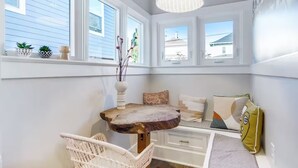 Une pièce lumineuse et bien éclairée, avec une grande fenêtre, une table en bois, une chaise blanche et un lit agrémenté d’oreillers à motifs.