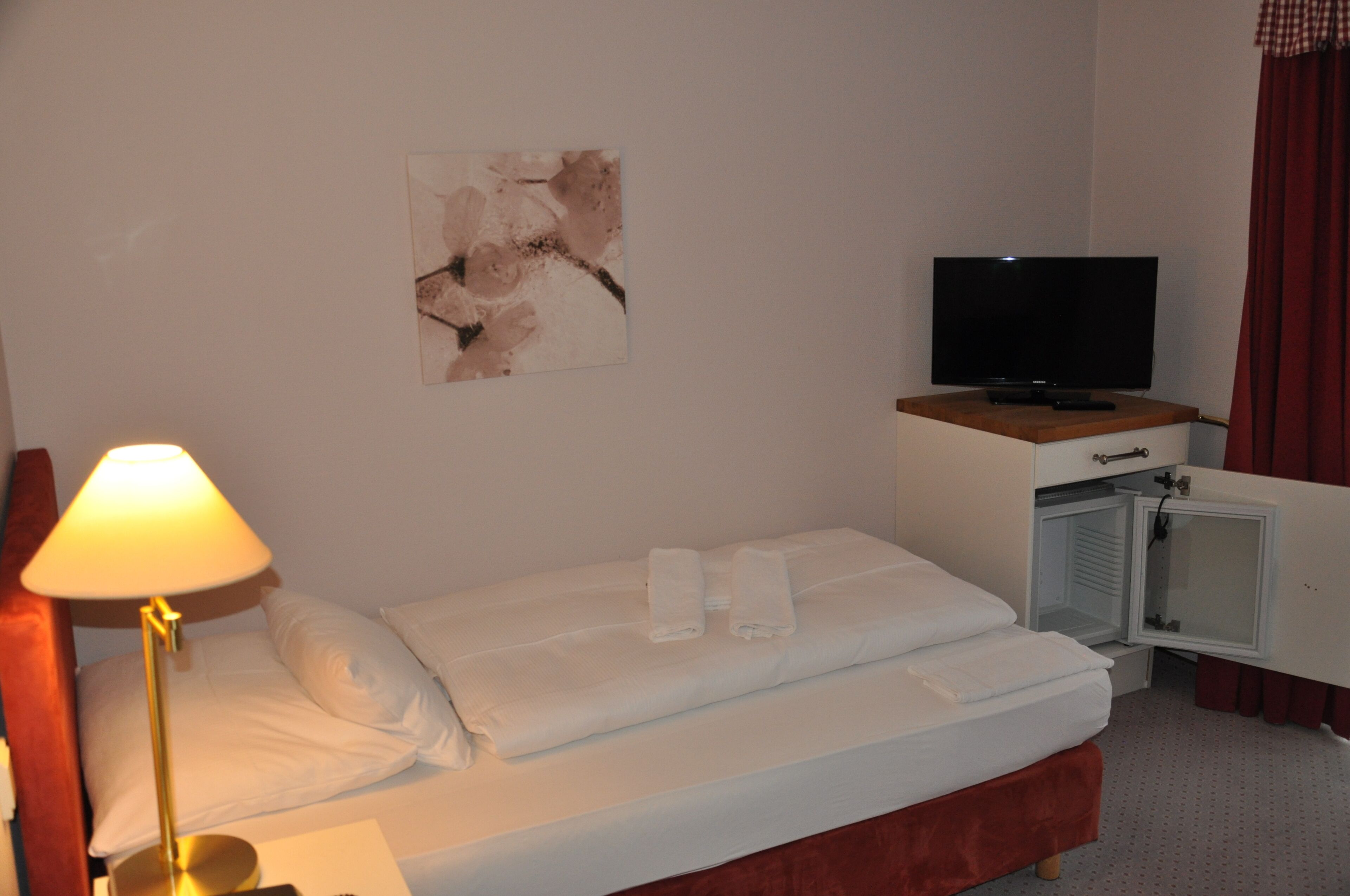 Una habitación de hotel con una cama, un televisor sobre un mueble, una lámpara y un armario pequeño.