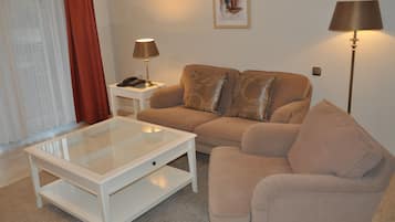 Una sala de estar con un sofá color crema, una mesa de centro de vidrio, una mesita de noche blanca con una lámpara y un cuadro en la pared.