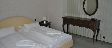 Una cama bien tendida con sábanas blancas, un aparador de madera, un espejo y una mesita de noche con una lámpara.