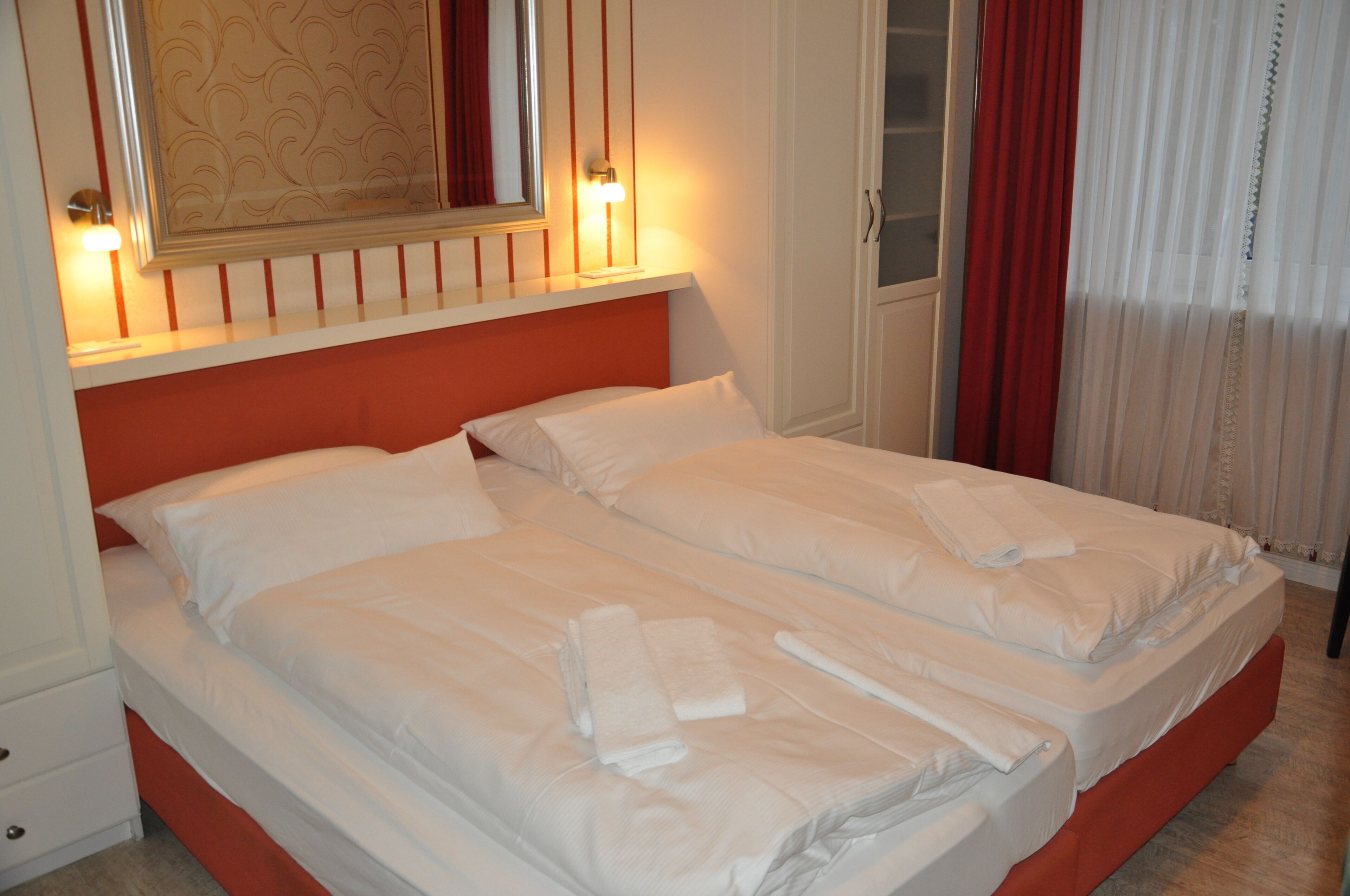A double bed with white linens and towels, a red headboard, and a mirror above it.