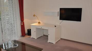A room with a white desk, a glass table, a gold desk lamp, a television on the wall, and a red curtain.