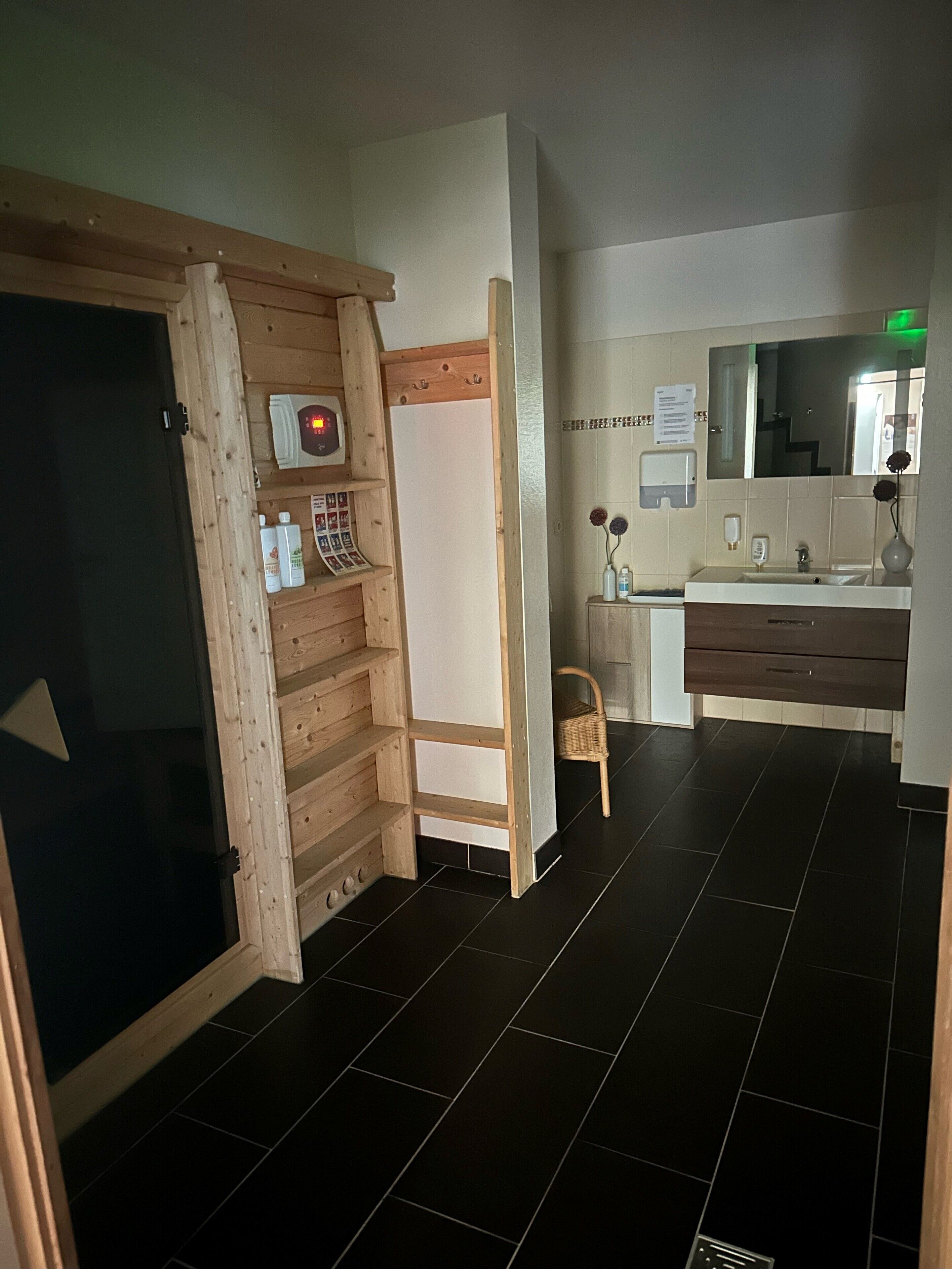 A hotel room with a wooden wardrobe, a whiteboard, a small desk with a chair, a bathroom with a mirror, and a black tiled floor.