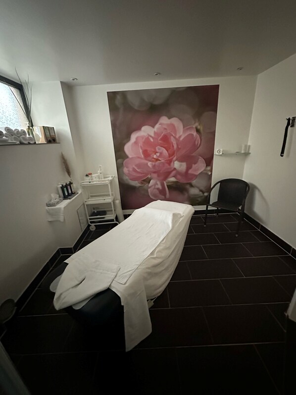 Um quarto clínico com uma cama de solteiro, uma cadeira, uma prateleira com artigos de higiene pessoal e um grande mural floral na parede.