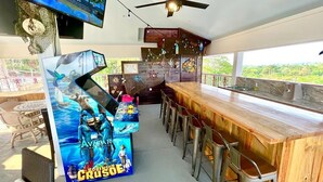 A bar area with a wooden counter and stools, a ceiling fan, and a TV mounted on the wall.