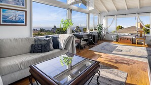 A spacious living area with a large window overlooking a scenic view, a comfortable sofa, a glass coffee table, and a desk with a globe. - Ocean View, Gym, Sauna, Garden, and Private Beach! (Laguna Beach)