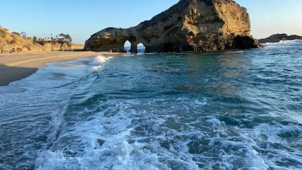 A coastal scene with a large rock formation featuring a natural archway, a sandy beach, and clear blue waters. - Ocean View, Gym, Sauna, Garden, and Private Beach! (Laguna Beach)