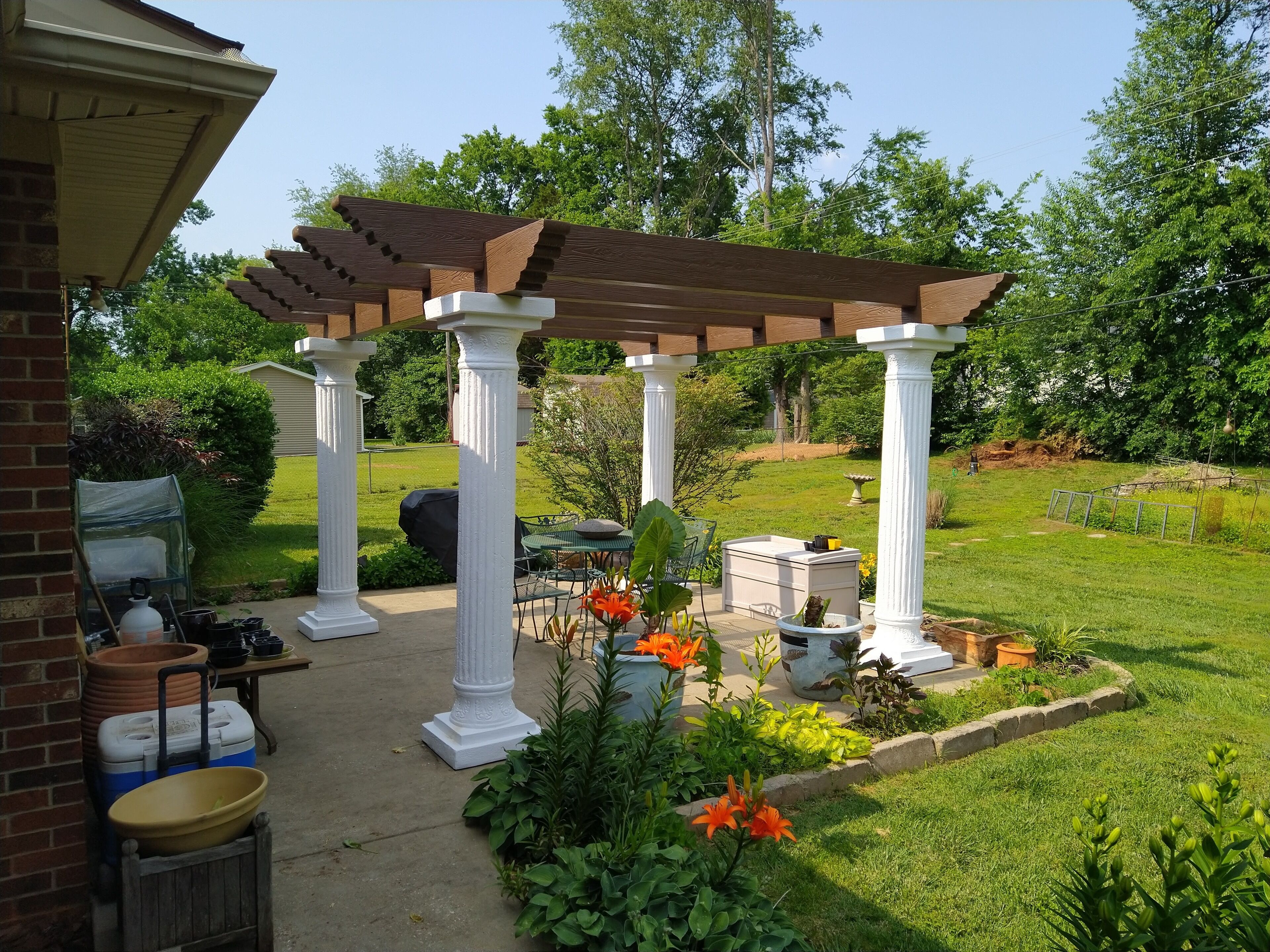 Un patio cubierto con columnas blancas, una pérgola, mobiliario exterior y un jardín con flores y plantas.