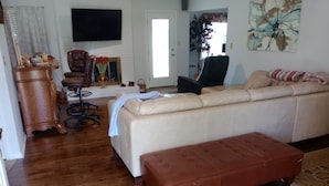 A living room with a beige sofa, a brown leather ottoman, a wooden cabinet, a television, and a painting on the wall.