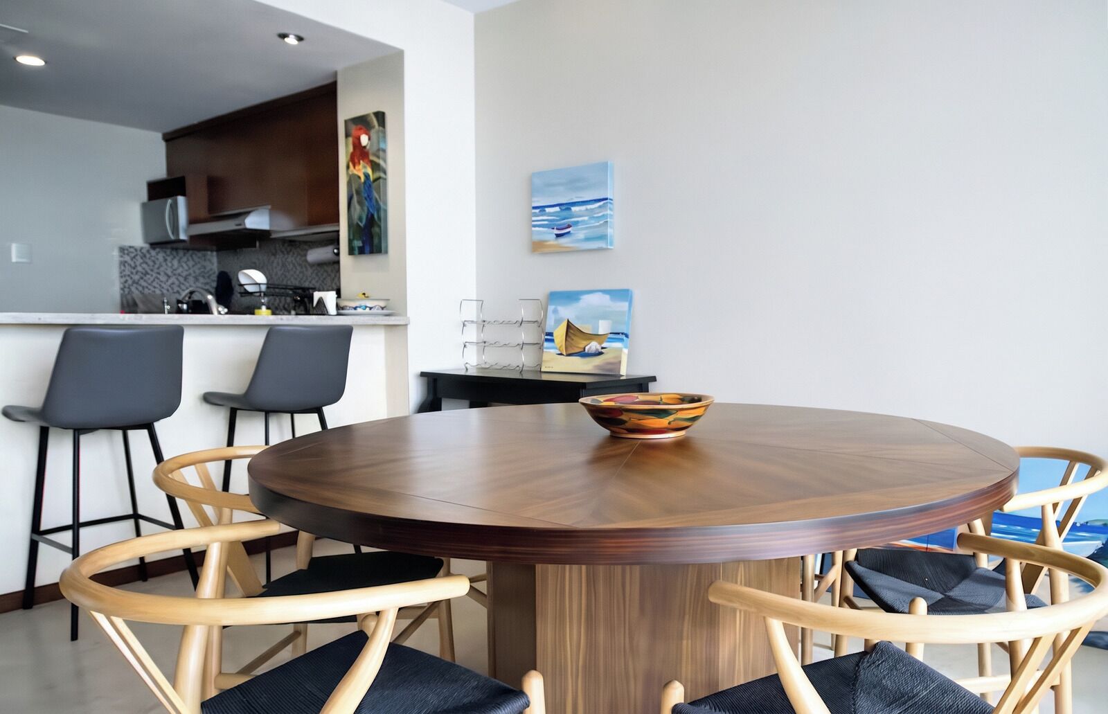 A round wooden dining table with a bowl, surrounded by four chairs with dark cushions.