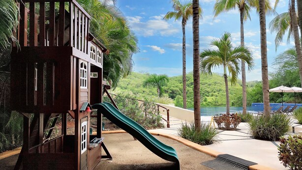 Un terrain de jeux avec un toboggan, entouré de palmiers et comprenant un espace piscine.