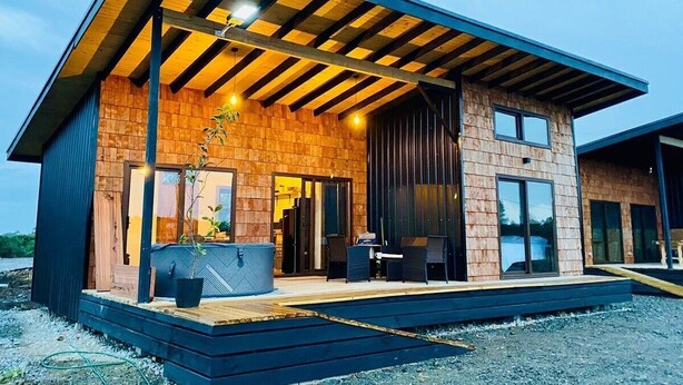 A modern two-story house with a wooden exterior, large windows, and a covered porch with outdoor seating. - Cabañas Lawal Pto Varas (Puerto Varas)