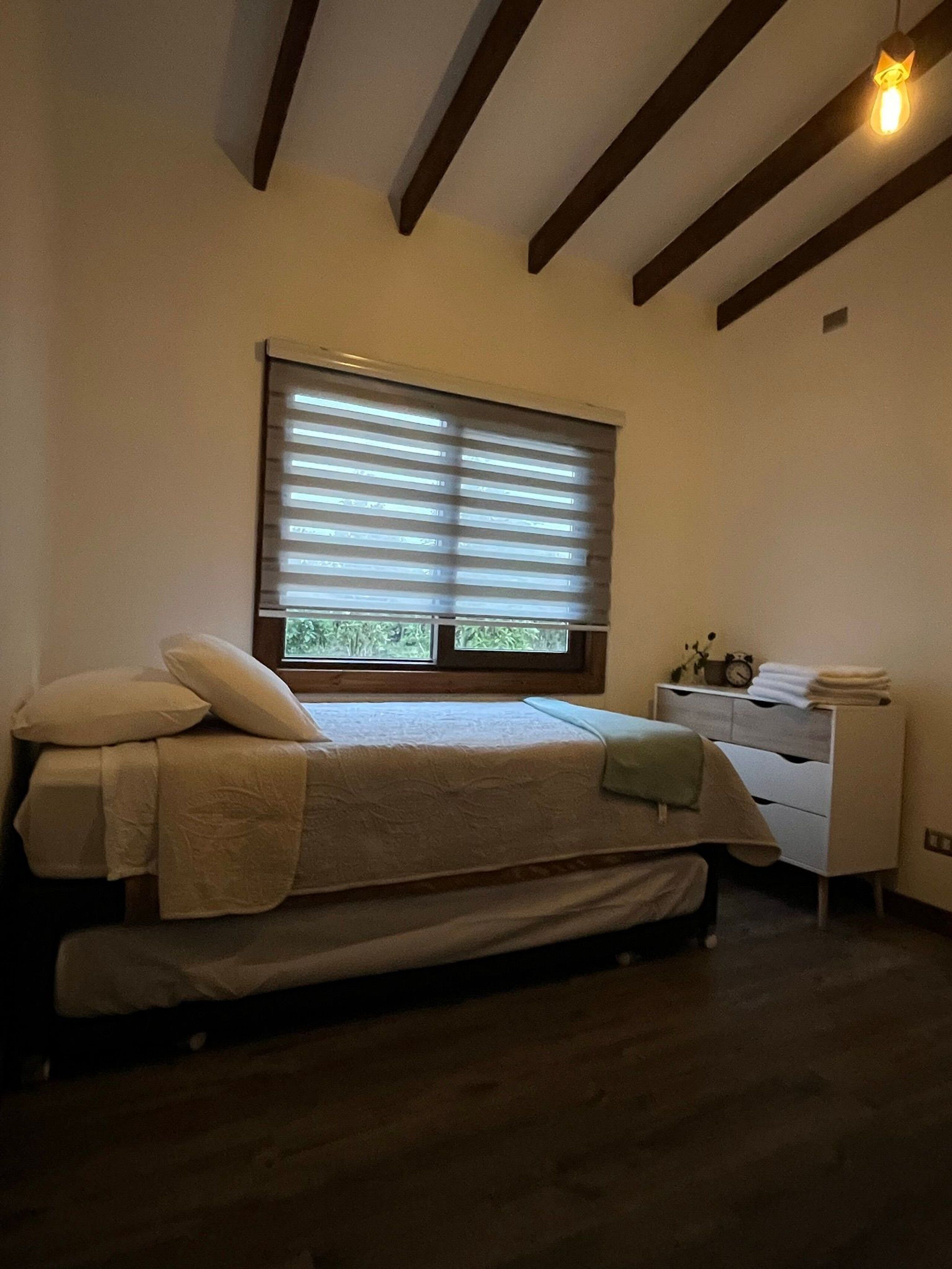 A bedroom with a bed, a window with blinds, a bedside table with a clock, and a small dresser.