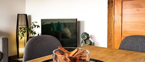 A wooden dining table with a glass vase containing wood shavings, a black wire basket, and a television in the background.
