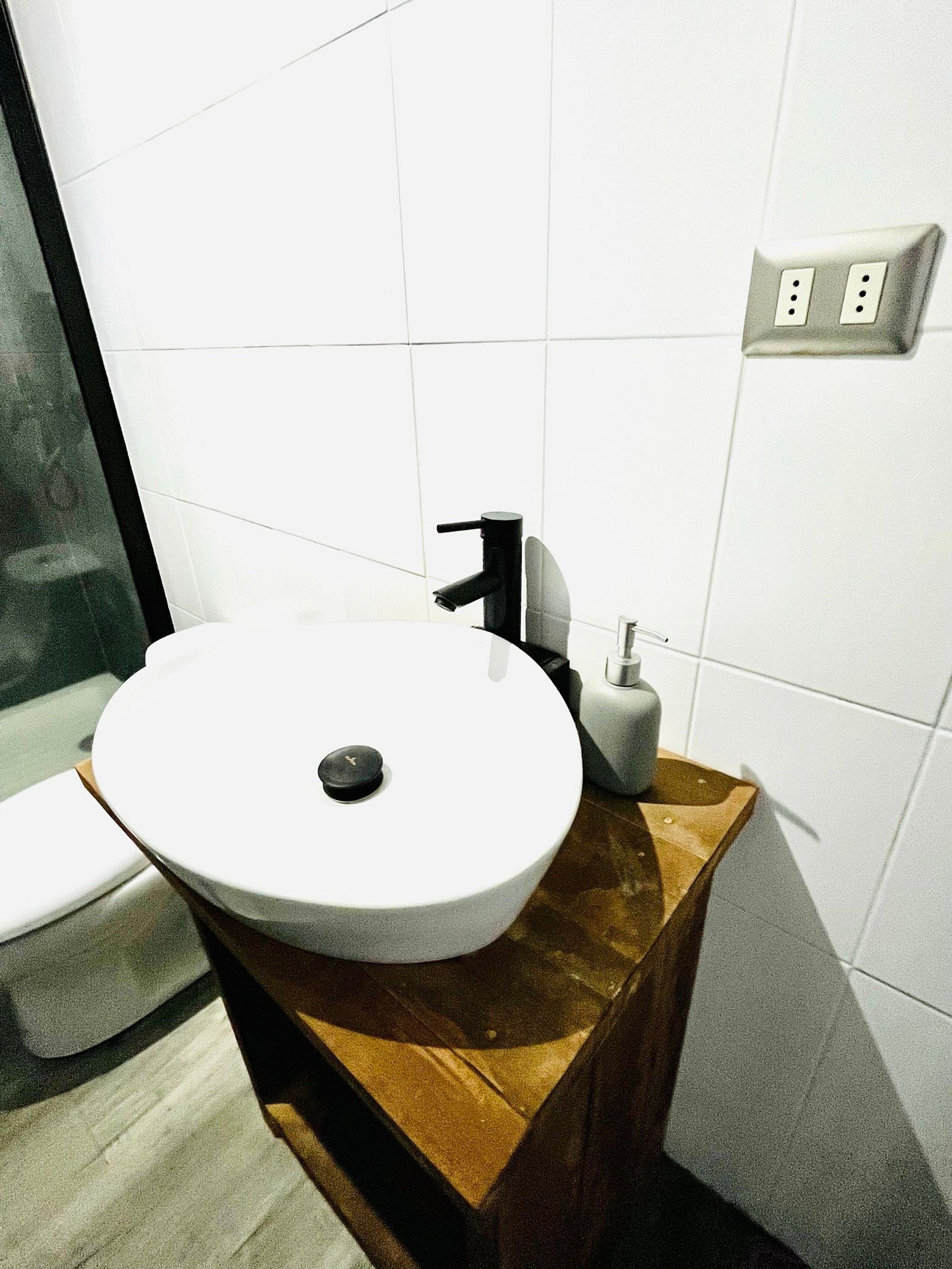 Un baño con un lavamanos blanco sobre un pedestal de madera, un grifo negro y un dispensador de jabón.