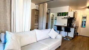 A modern living room with a white sofa, wooden flooring, and a kitchen area in the background.