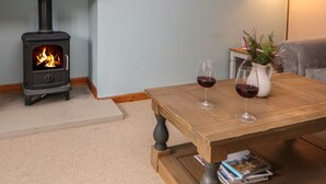 A cozy living room with a wood-burning stove, a wooden coffee table with wine glasses, a lamp, and a small plant. - Croft Cottage (Hope Valley)