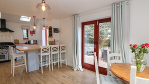 A modern kitchen with wooden flooring, white bar stools, a dining table with white chairs, and a view through a glass door to an outdoor seating area. - Croft Cottage (Hope Valley)