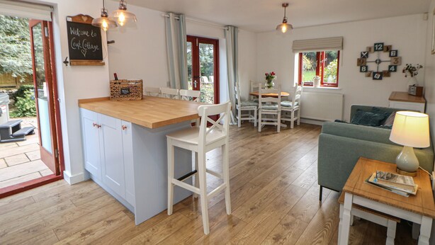 A cozy cottage with a kitchen, dining area, and living room. It features wooden flooring, white furniture, and a small island in the kitchen. - Croft Cottage (Hope Valley)