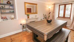 A dining area with a wooden table, a bowl, a bookshelf with books and decorative items, a mirror, and a window with curtains. - Croft Cottage (Hope Valley)