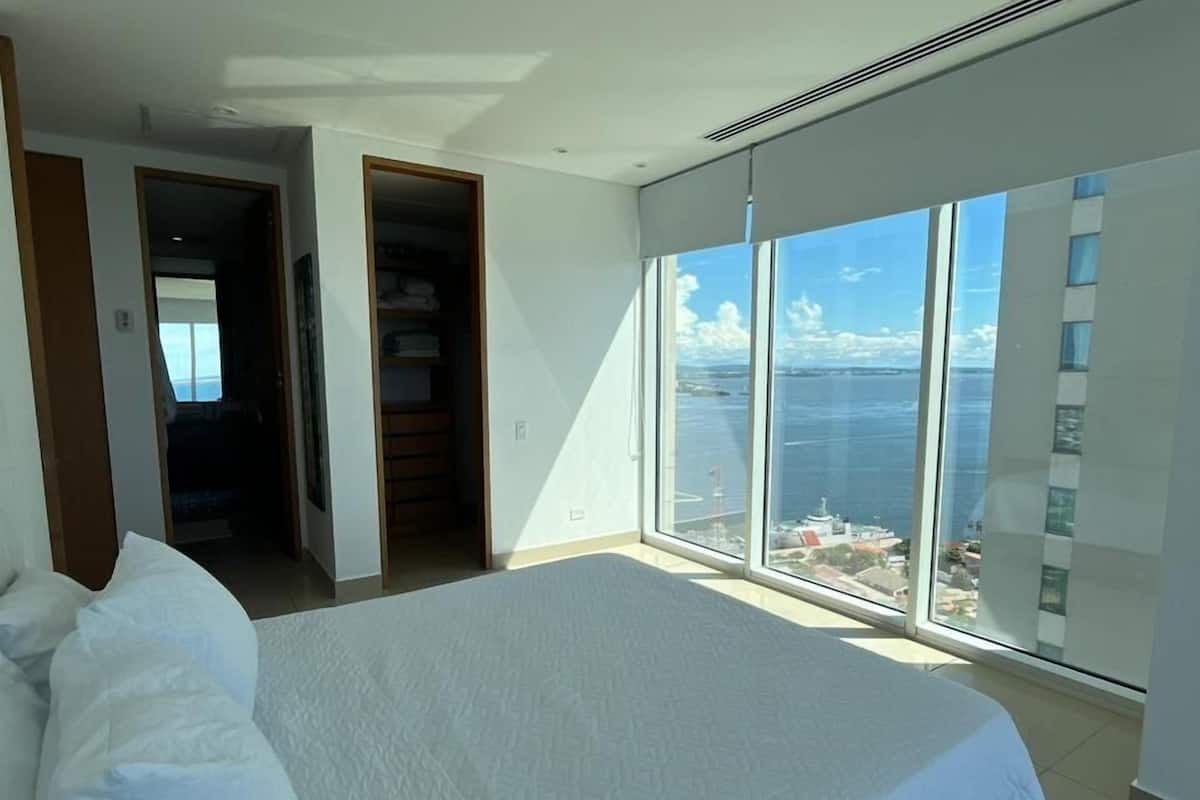 A bedroom with a large bed, a walk-in closet, and a view of the ocean through floor-to-ceiling windows.