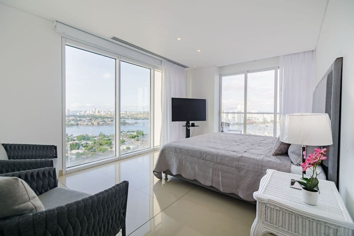 A modern hotel room with a large bed, a TV, and a view of a cityscape through large windows.