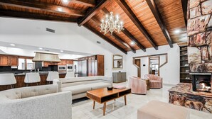 Un'ampia zona giorno con soffitto alto, travi in legno, un lampadario, un camino e una cucina moderna sullo sfondo.