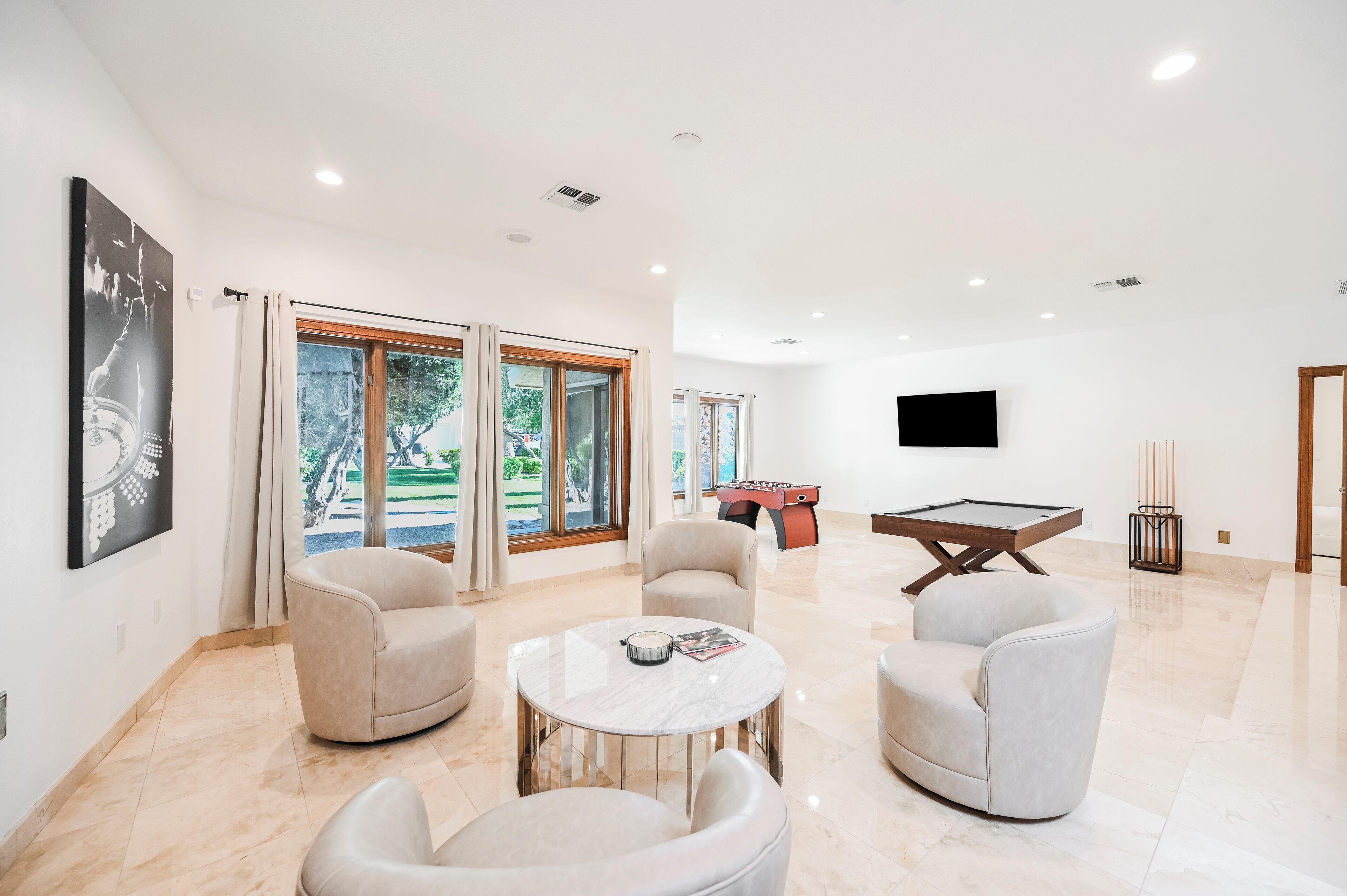 A modern living room with a pool table, comfortable seating, and large windows offering a view of the outdoors.