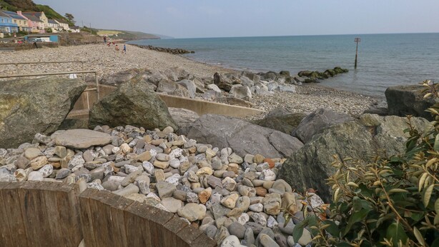 A rocky beach with a wooden walkway leading to the sea, houses in the background, and a clear sky. - The Stable (Narberth)