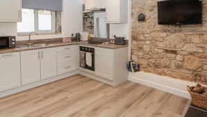 A modern kitchen with white cabinets, a stone wall, a flat-screen TV, and a wooden floor. - The Stable (Narberth)