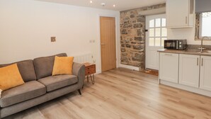 A modern living room with a grey sofa, a small wooden side table, and a kitchen area with white cabinets and a stone wall. - The Stable (Narberth)