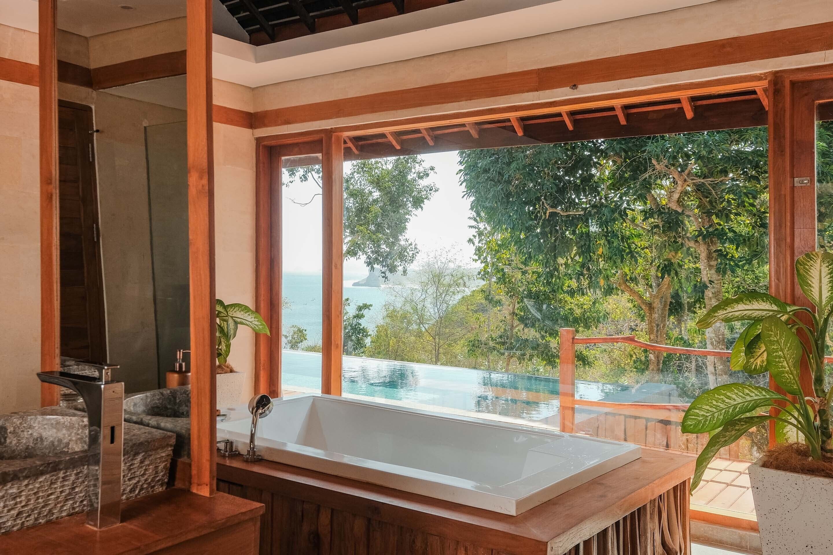 A modern bathroom with a large bathtub, wooden beams, and a view of a pool and greenery.