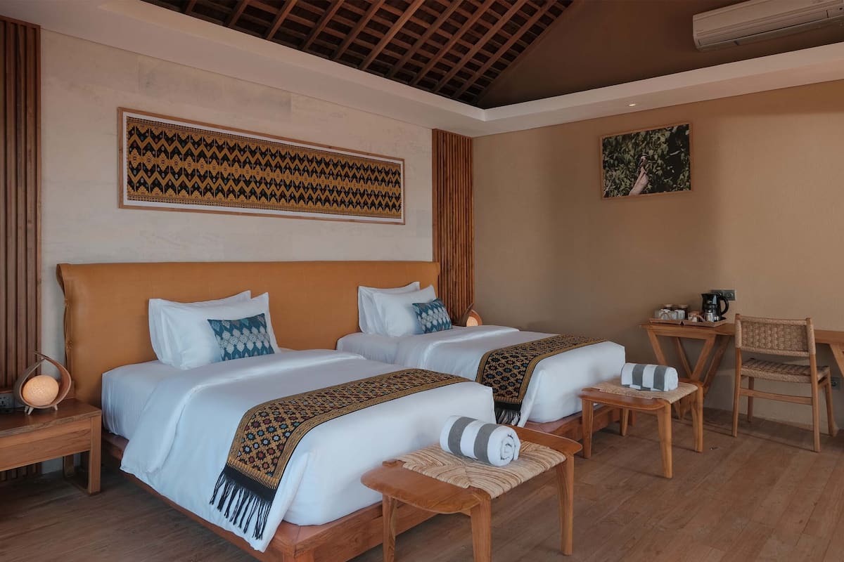 A hotel room with two beds, wooden furniture, a small table with a chair, and a wall-mounted air conditioner.