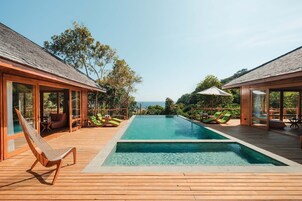 Zona de piscina con terrazas de madera y sillones de descanso, rodeada de muros de vidrio que ofrecen vistas al mar.