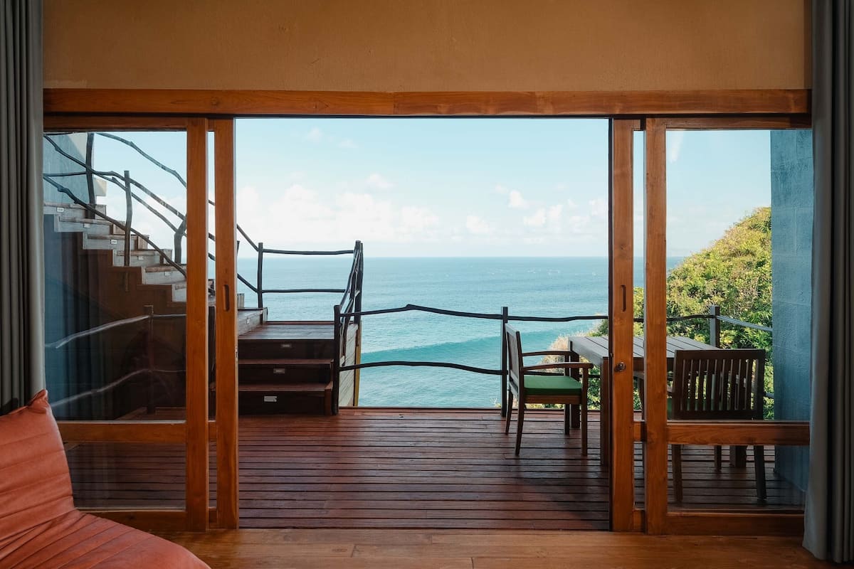 A room with a wooden deck leading to a balcony overlooking the sea.