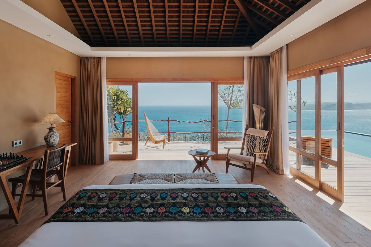 A bedroom with a large bed, a chessboard, a lamp, and a view of the sea through large windows.