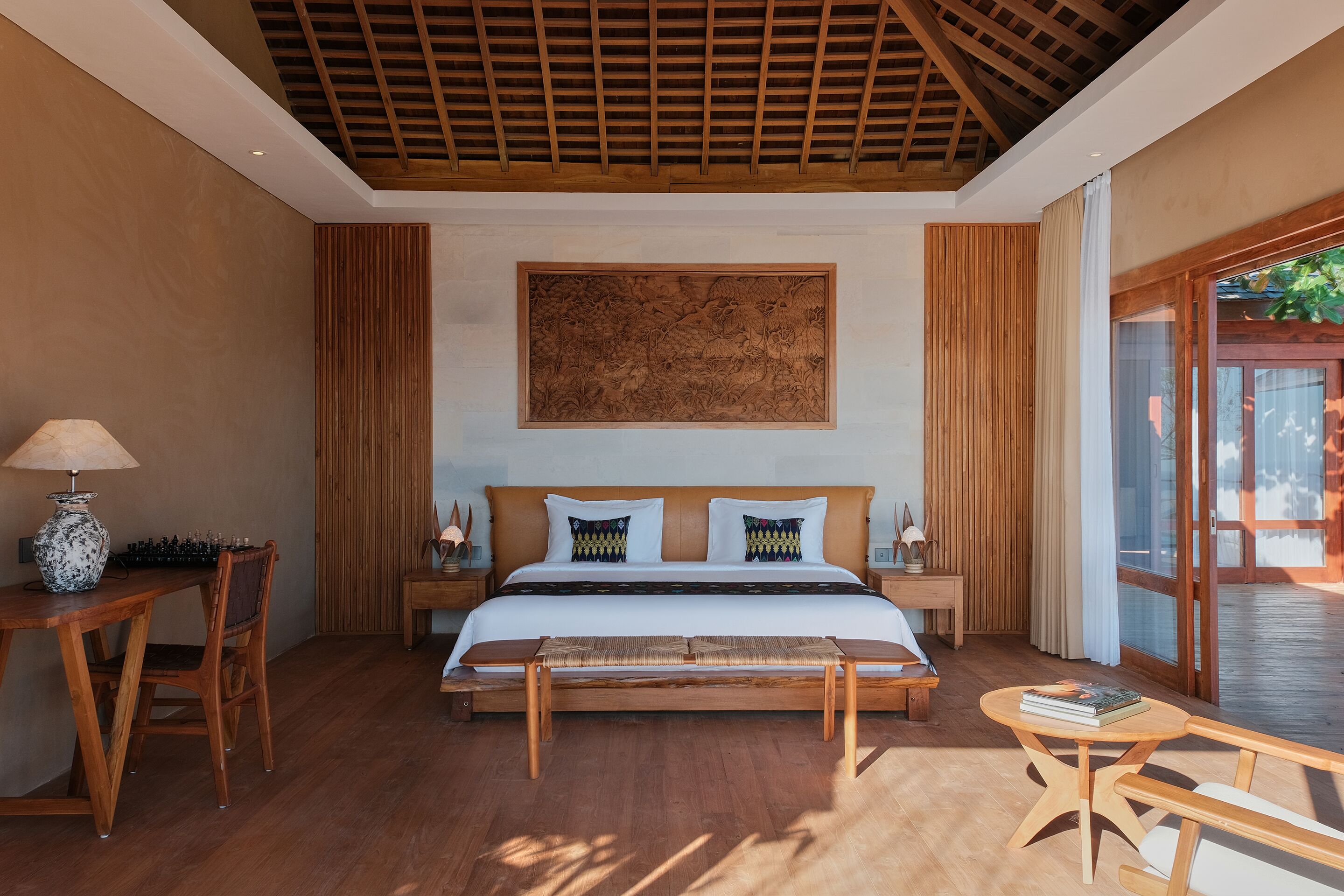 A bedroom with a wooden bed, a bench, a bedside table, and a large window.