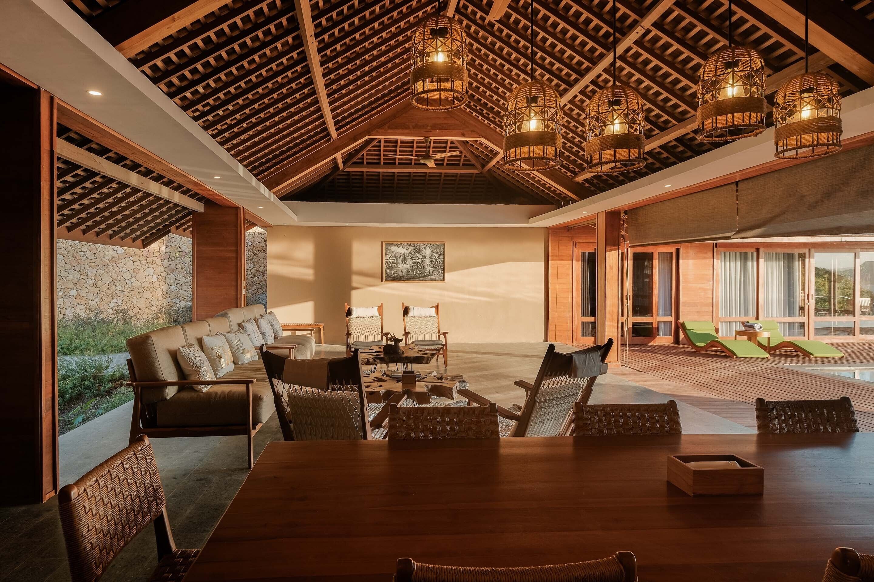 Un vaste espace de vie doté d’un plafond haut, de poutres en bois et d’une grande table à manger entourée de chaises. On y trouve également un coin salon avec un canapé et des fauteuils, ainsi qu’une vue sur une piscine et des chaises longues.