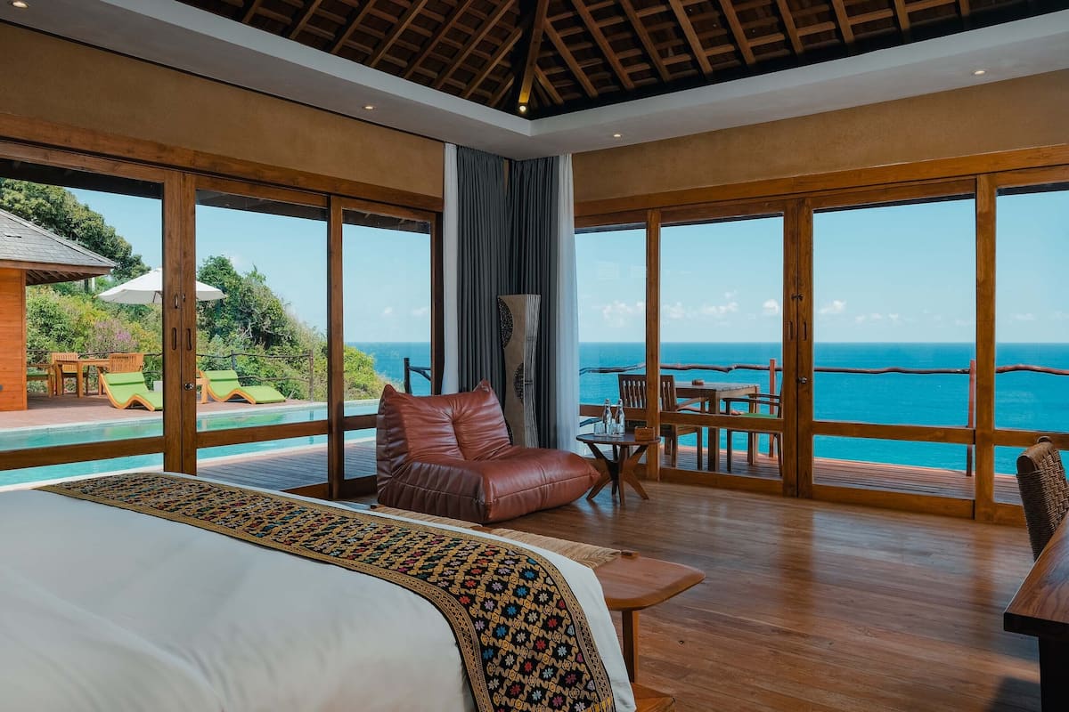 A spacious bedroom with a large bed, a leather armchair, a small table, and a view of the ocean through large windows.