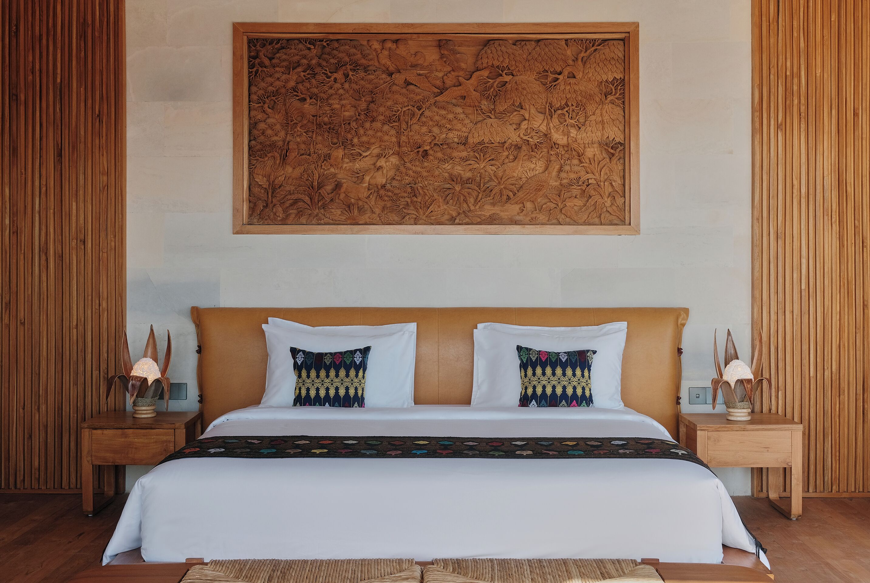 A bedroom with a large wooden headboard, a bed with white linens and patterned pillows, two bedside tables with lamps, and a large wooden artwork above the bed.