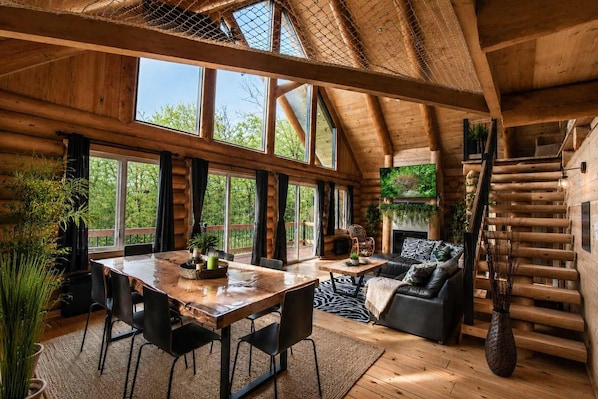 A log cabin interior with a dining area, sofa, fireplace, and large windows.