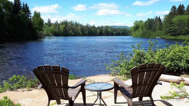 A lakeside view with two Adirondack chairs and a small table, surrounded by greenery and a clear sky.