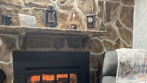 A stone fireplace with a lit fire, a grey armchair with a blanket, and a wall clock.