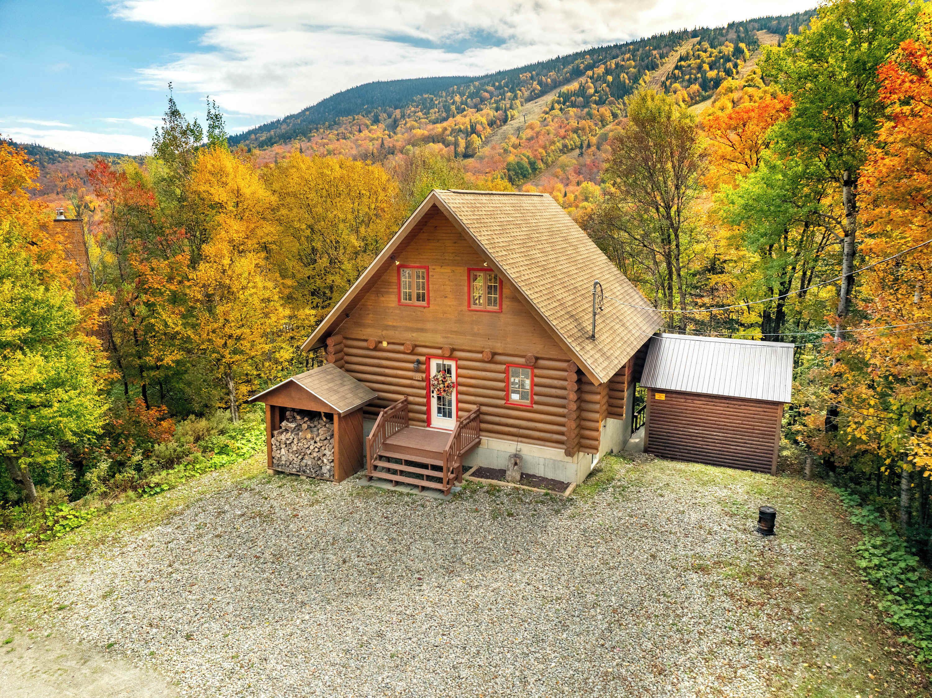 Eine Blockhütte mit Veranda und kleinem Schuppen, umgeben von Herbstlaub und einer Schotterauffahrt.